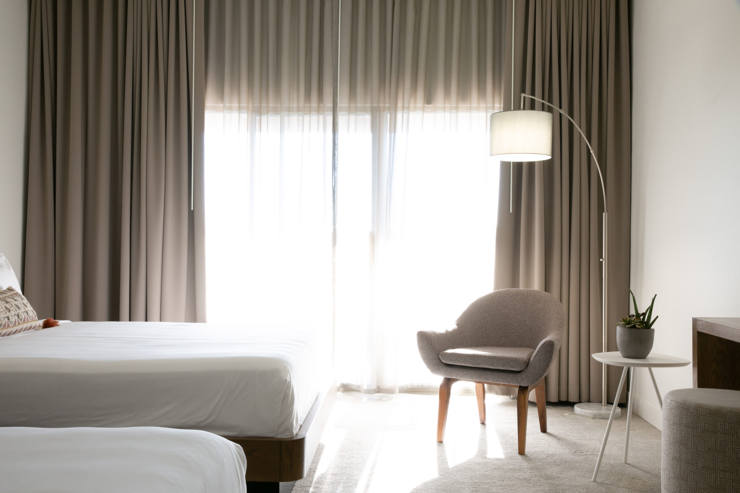 A wide view of a hotel room with a bed, chair, lamp, and window.